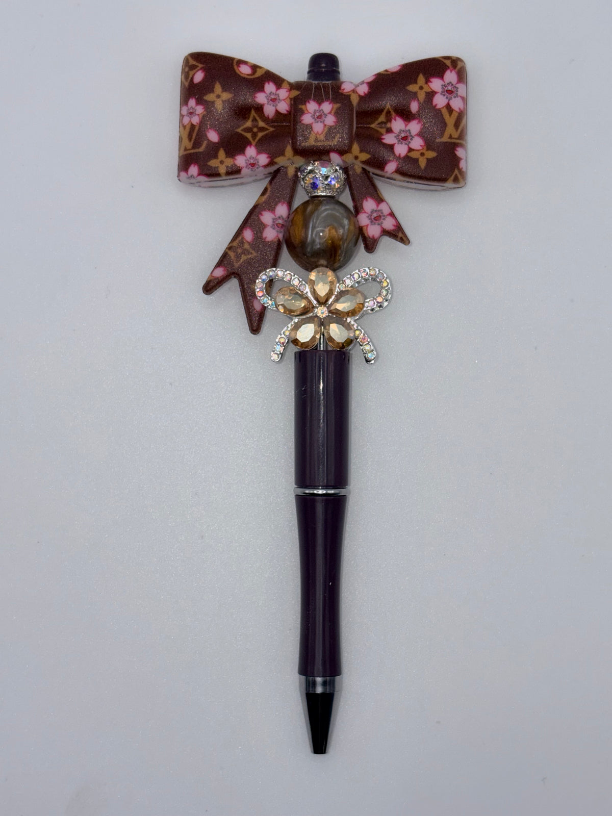 Dark beaded pen with a brown and pink floral patterned bow, brown swirled bead, and gold-tone sparkling butterfly accent on a plain background.