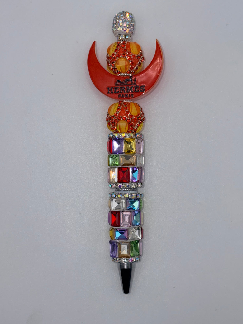 Beaded pen with orange crescent bearing Hermès Paris lettering, orange-and-yellow round beads, multicolor rectangular sparkling barrel accents, and dark tip.