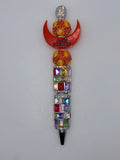 Beaded pen with orange crescent bearing Hermès Paris lettering, orange-and-yellow round beads, multicolor rectangular sparkling barrel accents, and dark tip.
