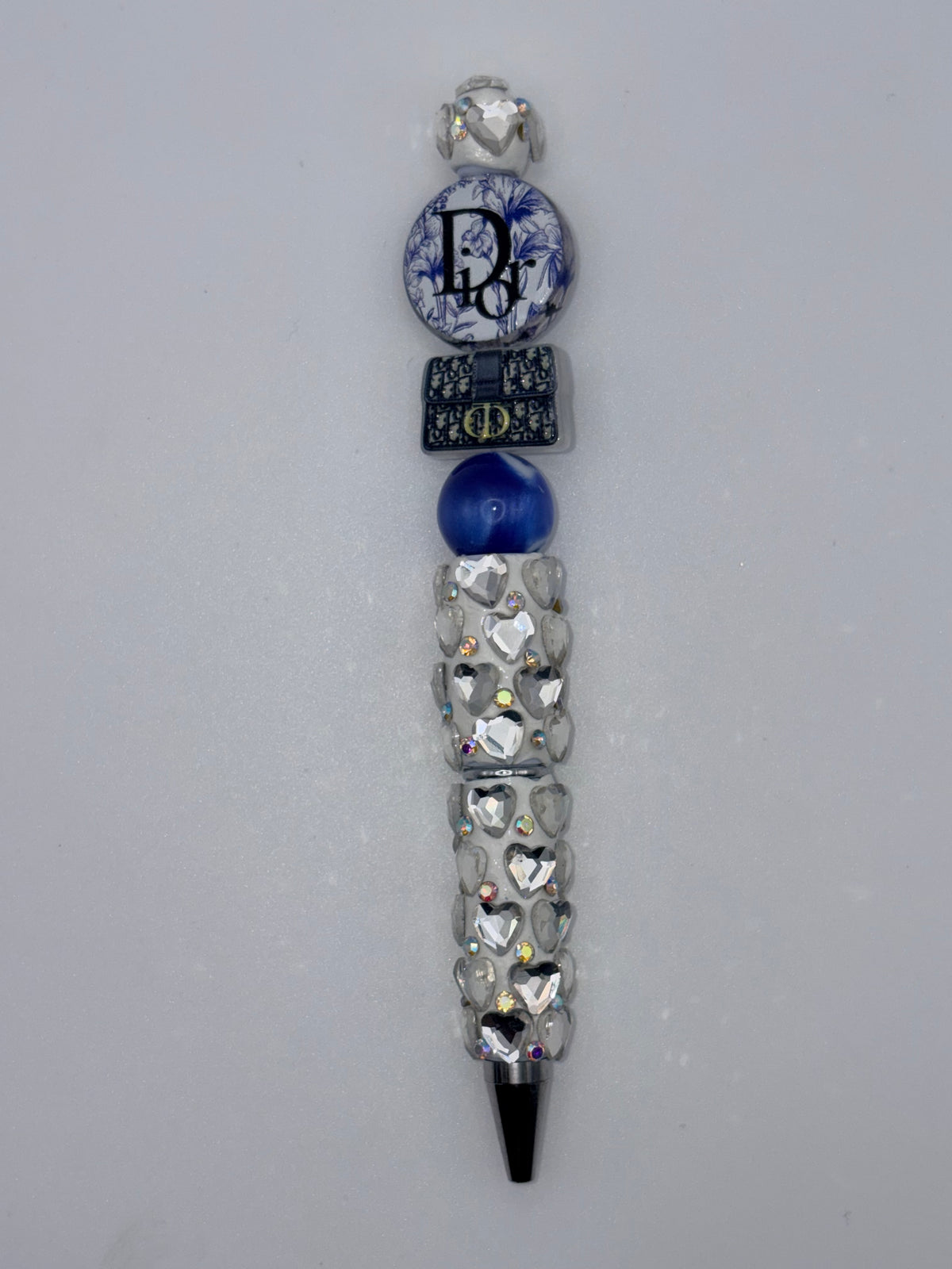 Blue and silver beaded pen with reflective heart-decorated barrel, deep blue bead, navy handbag-shaped accent, and round blue-and-white floral lettered accent.