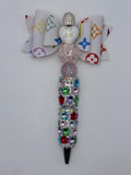 Beaded pen with white multicolor monogram-patterned bow, white heart accent, pale pink flower and glitter bead, multicolor sparkling barrel, and silver-tone tip.