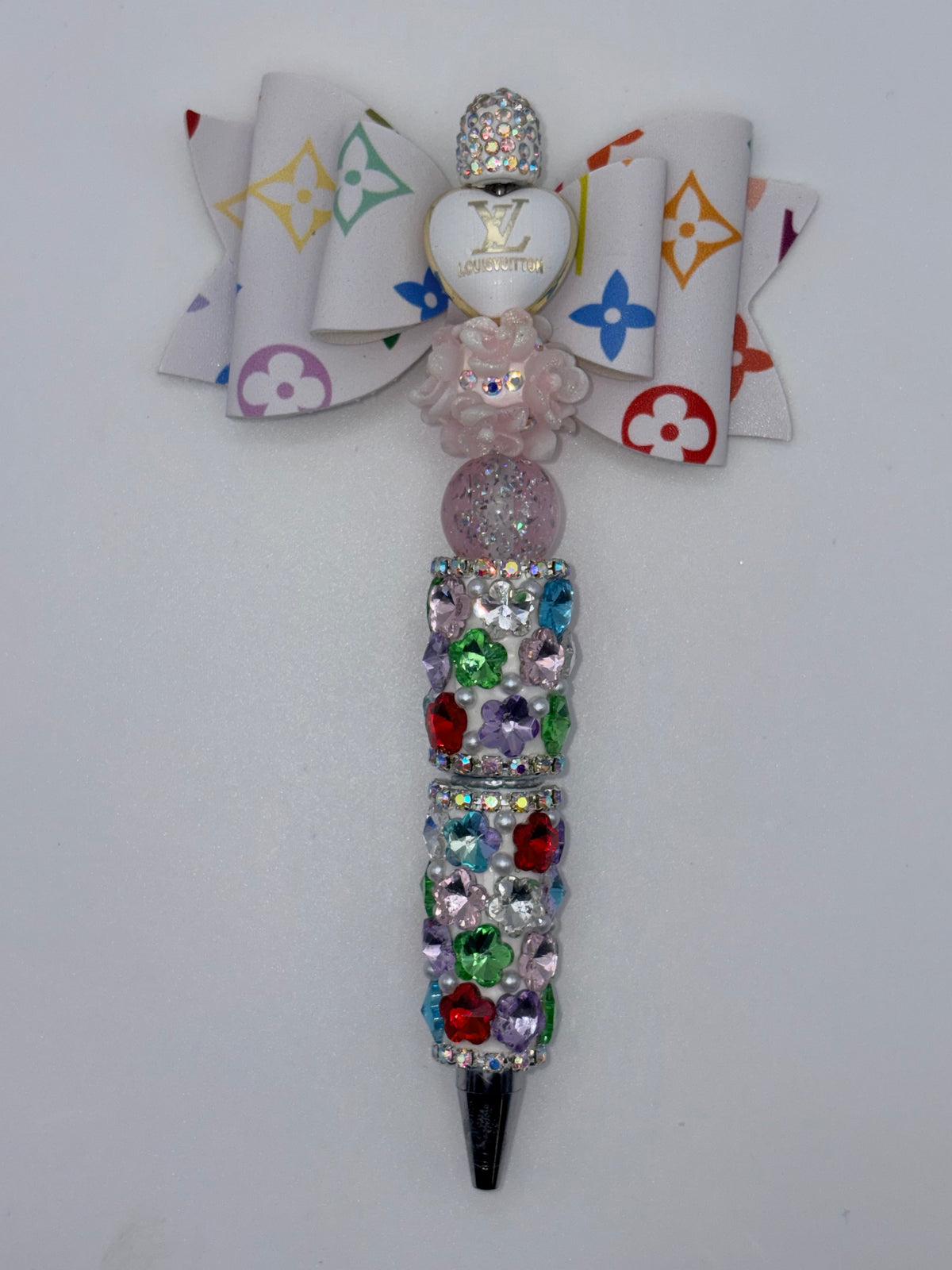 Beaded pen with white multicolor monogram-patterned bow, white heart accent, pale pink flower and glitter bead, multicolor sparkling barrel, and silver-tone tip.
