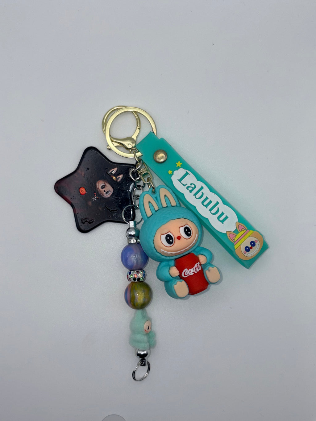 Teal Labubu-themed wristlet keychain with bunny-eared character holding a red soda can, black star charm, colorful beaded dangle, and gold-tone ring.