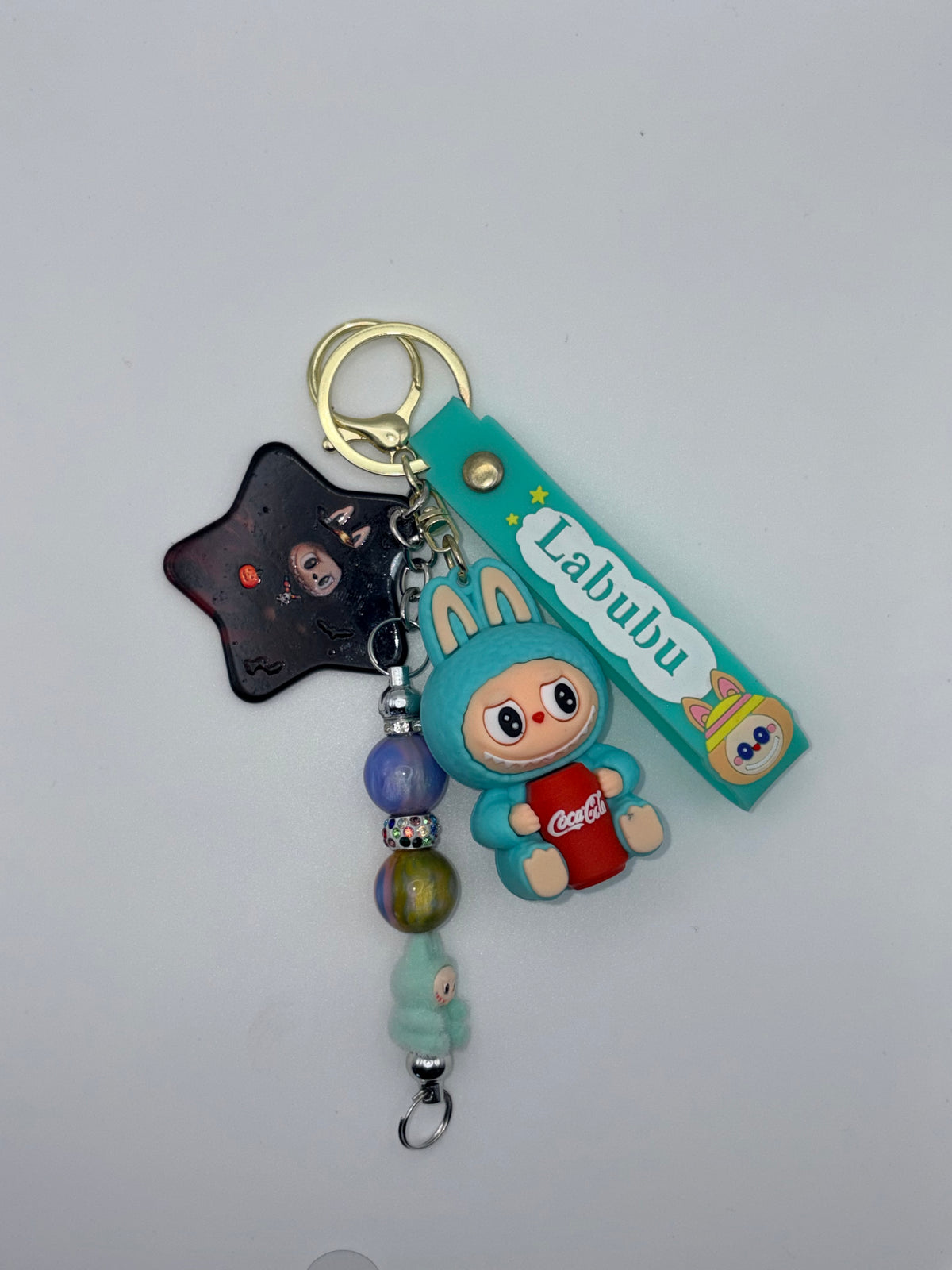 Teal Labubu-themed wristlet keychain with bunny-eared character holding a red soda can, black star charm, colorful beaded dangle, and gold-tone ring.